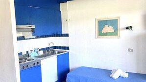 A small kitchenette with a stove, sink, and blue cabinets. There is a bed with a blue bedspread and a framed picture on the wall. - P.Rotondo1 M131 Two-Bedroom Apartment 5 SEA VIEW (Porto Rotondo)