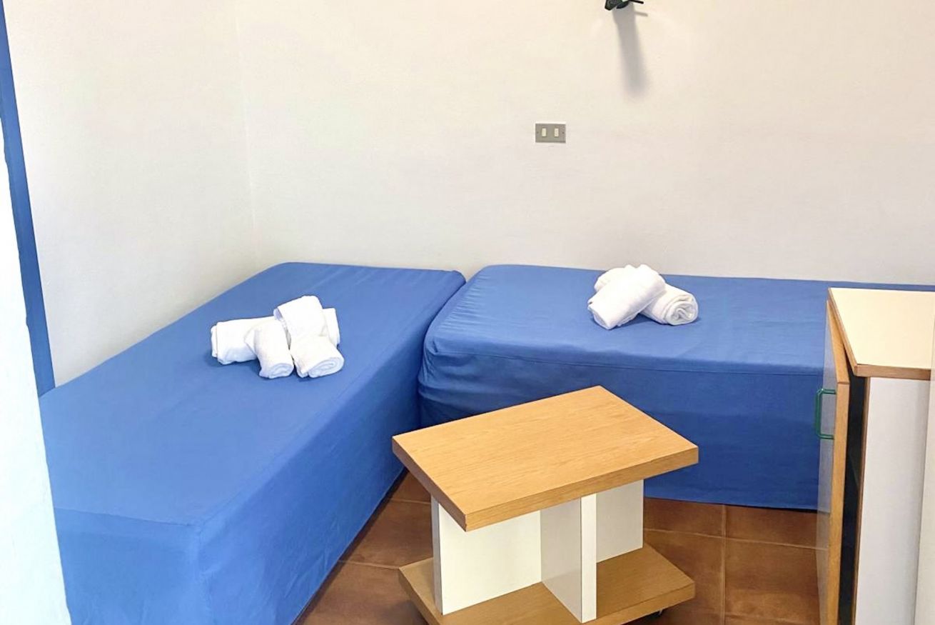 Une petite pièce avec deux lits, chacun recouvert d’un drap bleu et d’une serviette blanche. Une petite table en bois se trouve au centre de la pièce.