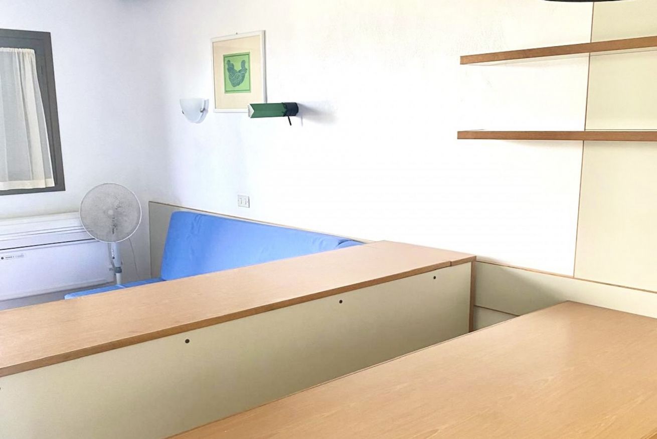 Une pièce comprenant un lit, un bureau, une chaise, un ventilateur fixé au mur et un tableau encadré.