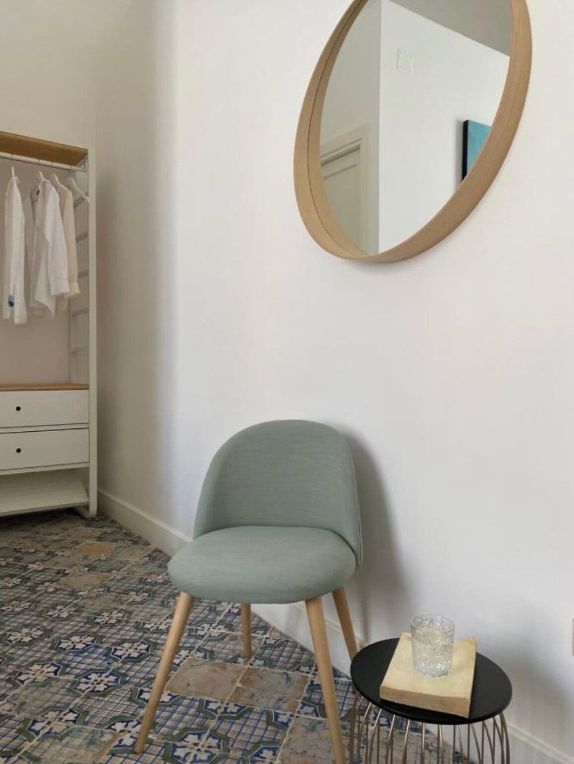A room with a chair, a small table with a glass, a mirror on the wall, a wardrobe, and patterned floor tiles.