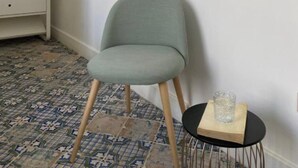 A room with a chair, a small table with a glass, a mirror on the wall, a wardrobe, and patterned floor tiles.