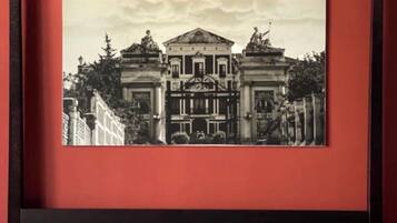 Une photographie en noir et blanc, encadrée, représentant un bâtiment classique avec des colonnes, est accrochée au-dessus d’un lit recouvert d’une courtepointe blanche et agrémenté de deux oreillers rouges.