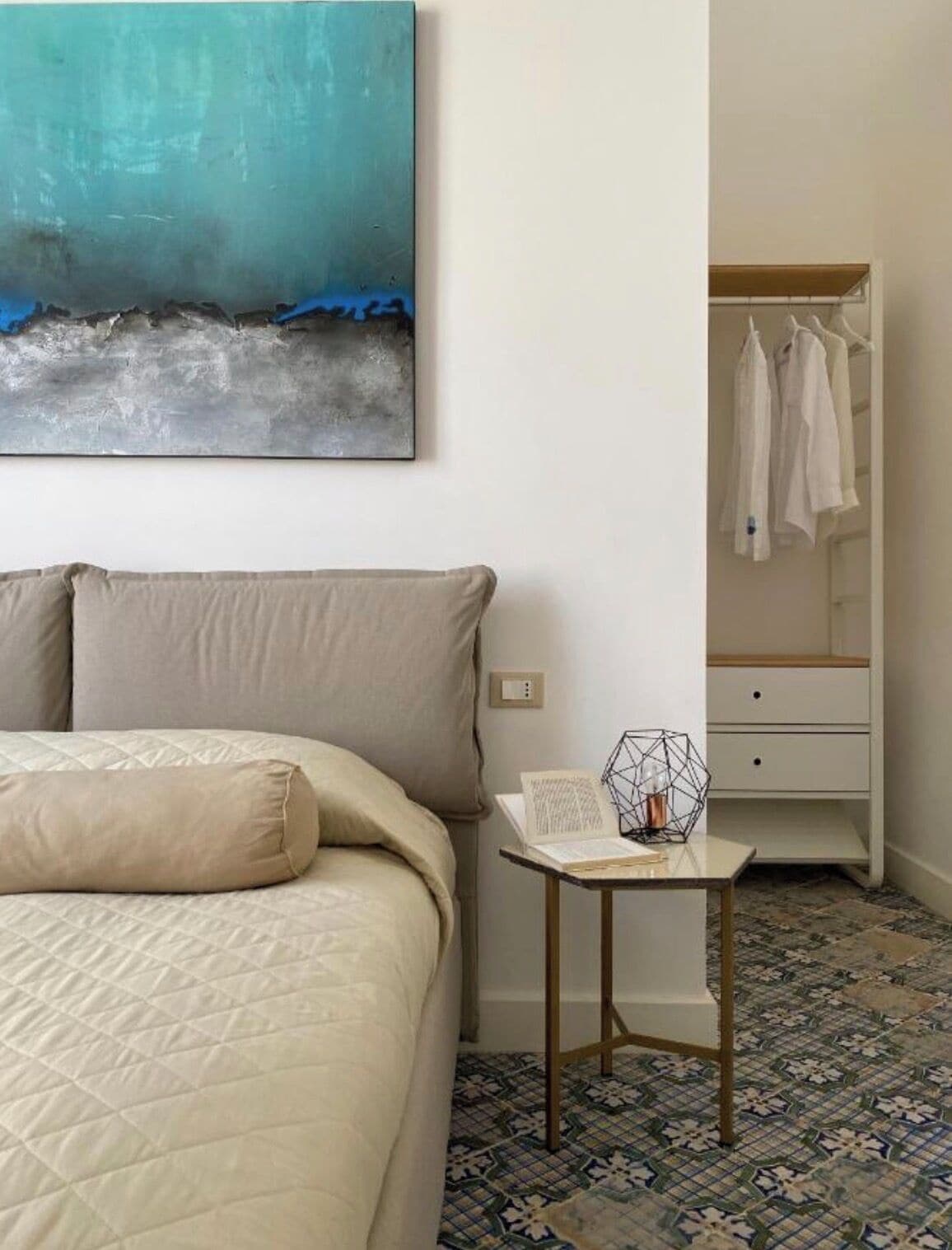 A hotel room with a bed, a small table with a book and a geometric lamp, a wardrobe, and a large abstract painting on the wall.