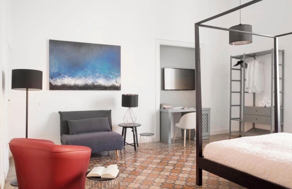 A modern hotel room with a bed, a desk, a chair, a sofa, a red armchair, a painting, and a patterned floor.