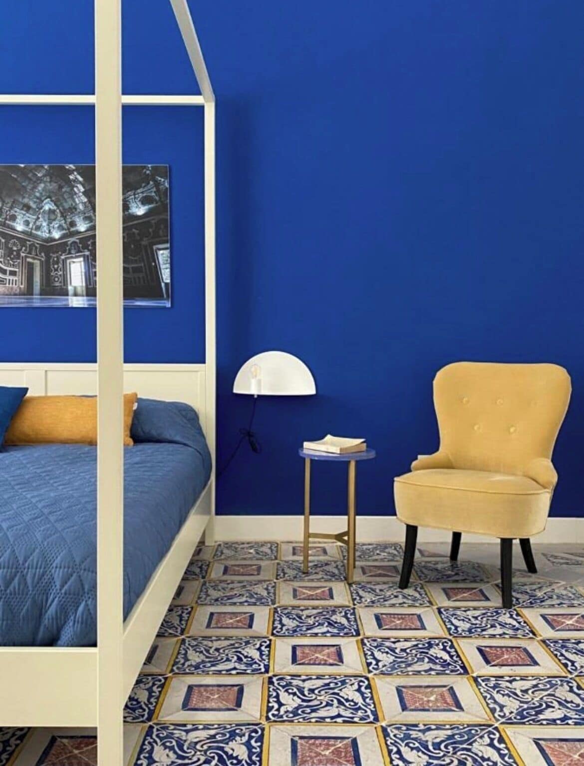 A bedroom with a four-poster bed, a small round table, a yellow armchair, and a patterned floor.