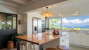 A modern kitchen with a large island, bar stools, a hanging light fixture, and a view of the ocean.