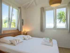 A bedroom with a bed, two windows, a lamp, and a view of greenery outside. - Villa Bananes Rouges (Trois-Rivières)