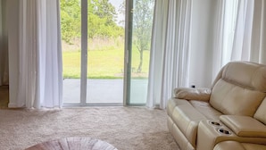 A room with a beige leather sofa, a carpeted floor, and a sliding glass door leading to a grassy outdoor area. - Cozy 2-bedroom cottage with AC in cool Jacksonville 55+ community in Mandarin (Jacksonville)