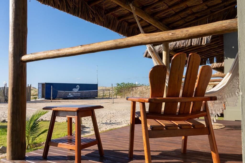 A wooden deck with a chair and table, a thatched roof, and a view of the beach and shipping containers in the background.