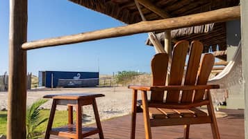 A wooden deck with a chair and table, a thatched roof, and a view of the beach and shipping containers in the background.