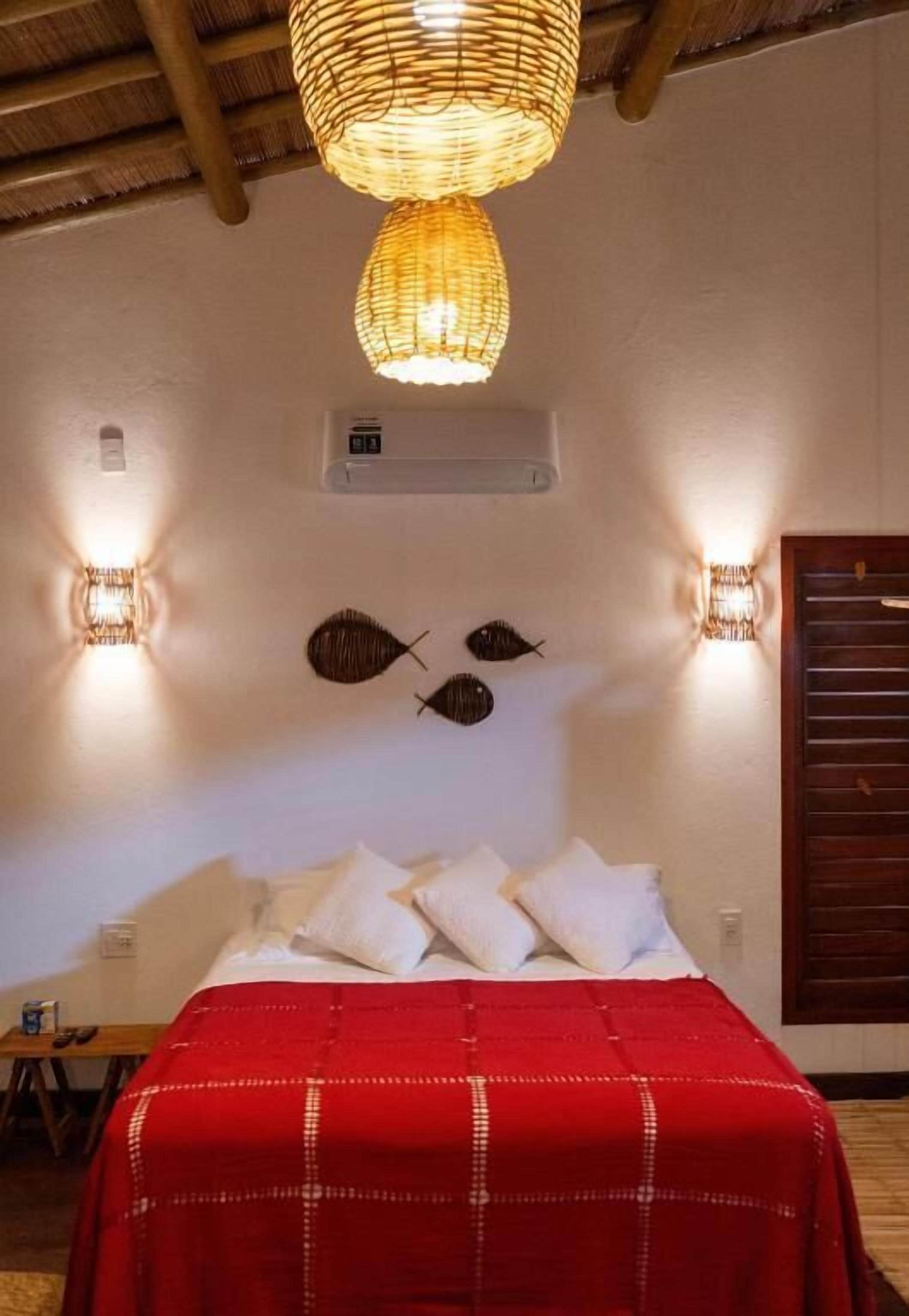 A bedroom with a bed, a red bedspread, wall-mounted lights, and a woven ceiling light fixture.