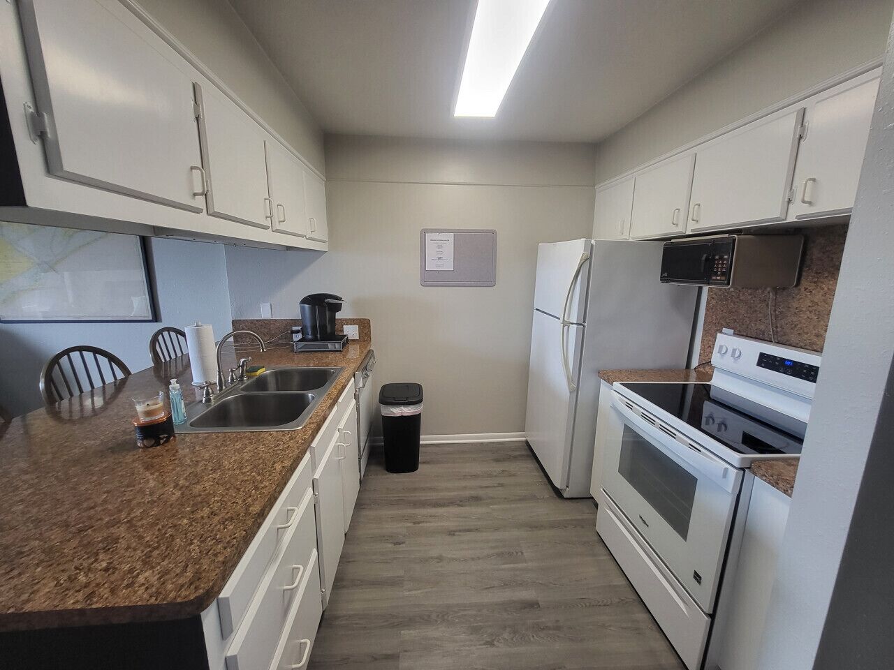 A compact kitchen with white cabinets, a granite countertop, stainless steel appliances, and a trash bin.