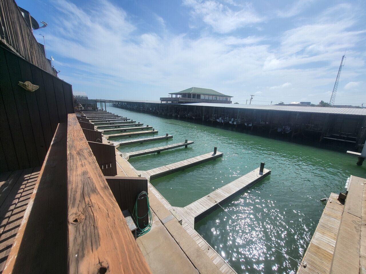 Vista al agua con una terraza de madera, un muelle que se adentra en el agua y un edificio con techo verde al fondo.