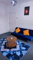 Sala de estar con un sofá azul, dos cojines naranja, una mesa de centro redonda de madera con un jarrón de flores, una alfombra estampada en azul y negro, una puerta blanca y un cuadro abstracto colgado en la pared.