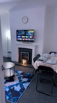 Una habitación con chimenea, un televisor montado sobre ella, una mesa de comedor con platos y vasos, y un reloj en la pared.
