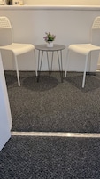 Una pequeña mesa redonda con una planta en maceta, dos sillas blancas con respaldos perforados y un suelo alfombrado.