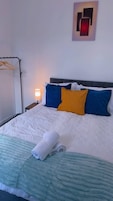 Una cama bien hecha con un edredón blanco acolchado, dos almohadas en azul y amarillo, y una toalla doblada sobre la cama.