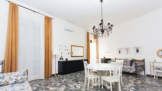 Uma sala de estar com uma mesa de jantar, cadeiras, sofá, lustre e uma janela grande com cortinas.