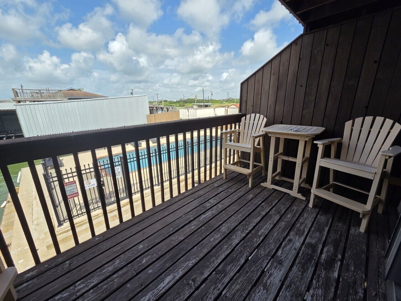 Terraza de madera con dos sillas Adirondack y una mesita, con vista a un área cercada que tiene una piscina.
