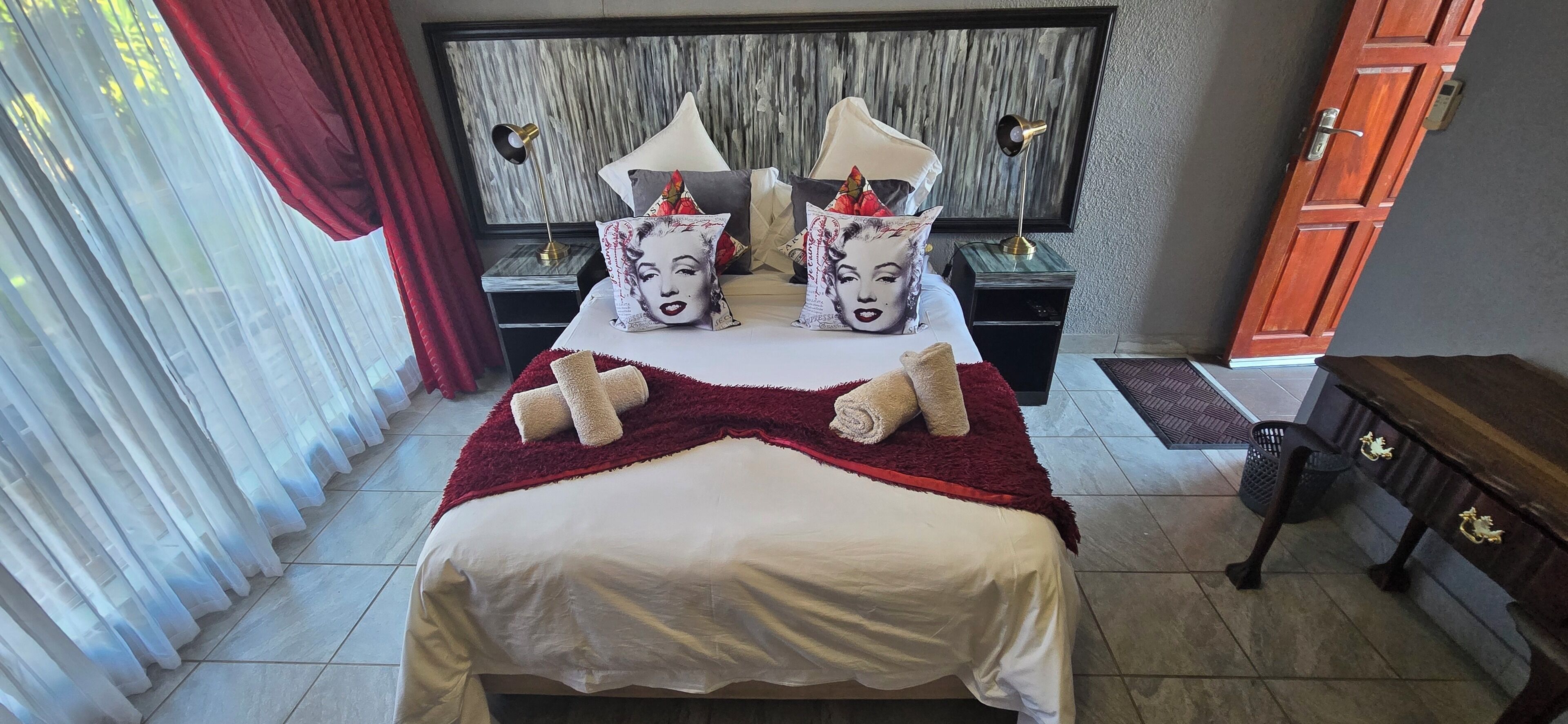 A hotel room with a bed, pillows, a bedside table, a lamp, a nightstand, a wooden bench, and a door.