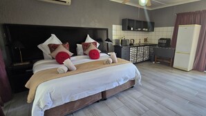 A bedroom with a large bed, a bedside table, a TV, and a small kitchenette with a fridge, microwave, and stove.