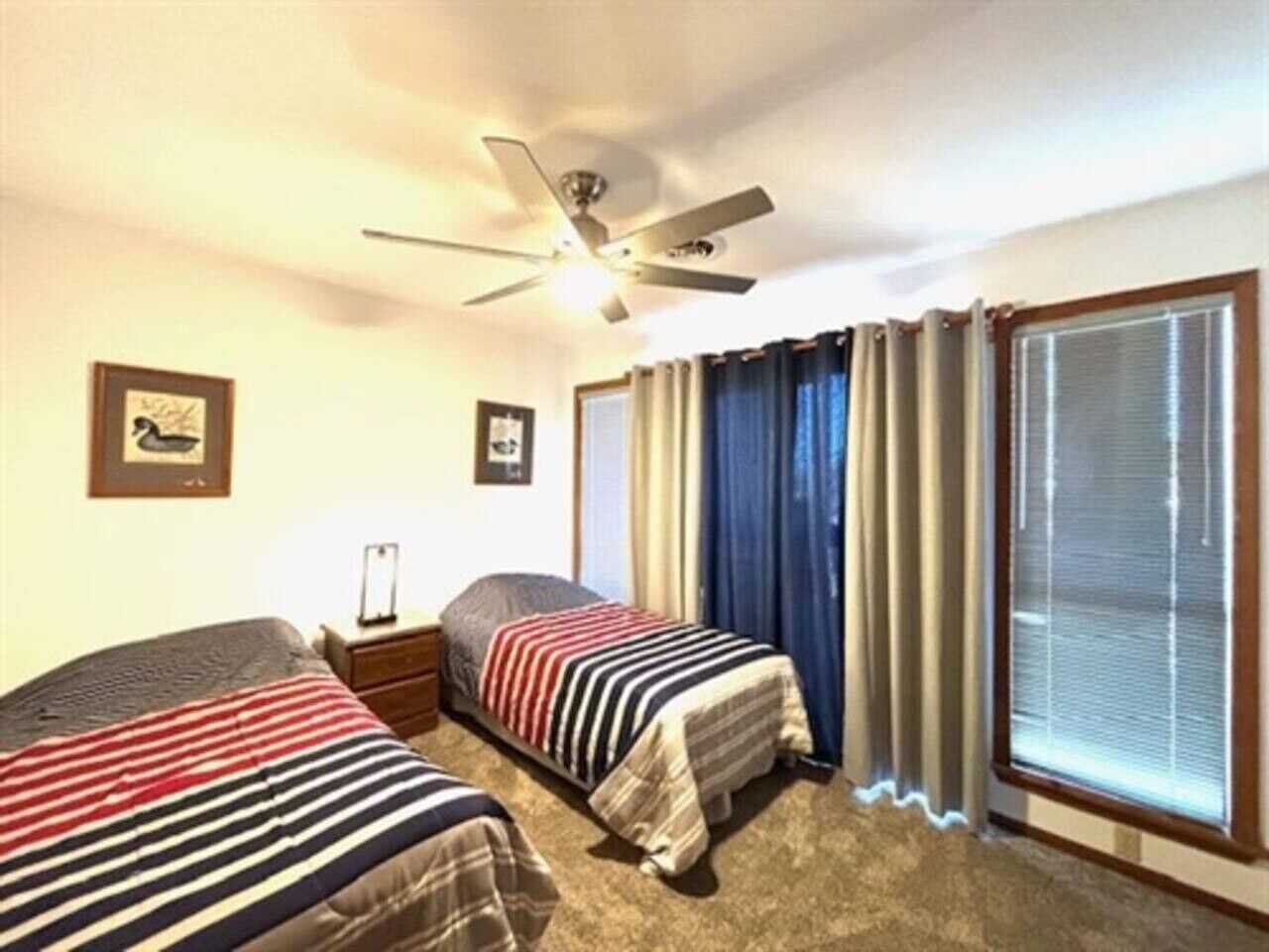Camera d'albergo con due letti, una ventola a soffitto, una finestra con tende e un quadro appeso al muro.