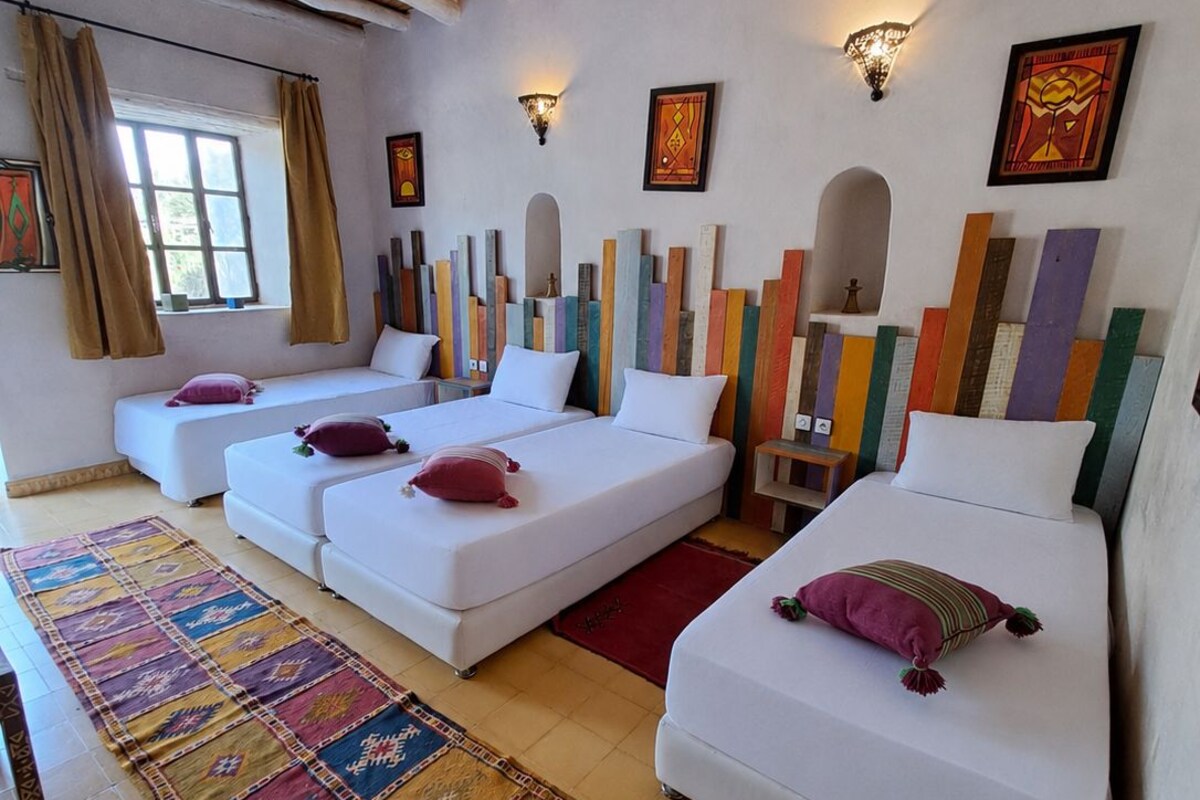 A hotel room with a thatched roof, white beds, colorful artwork, and a patterned rug.