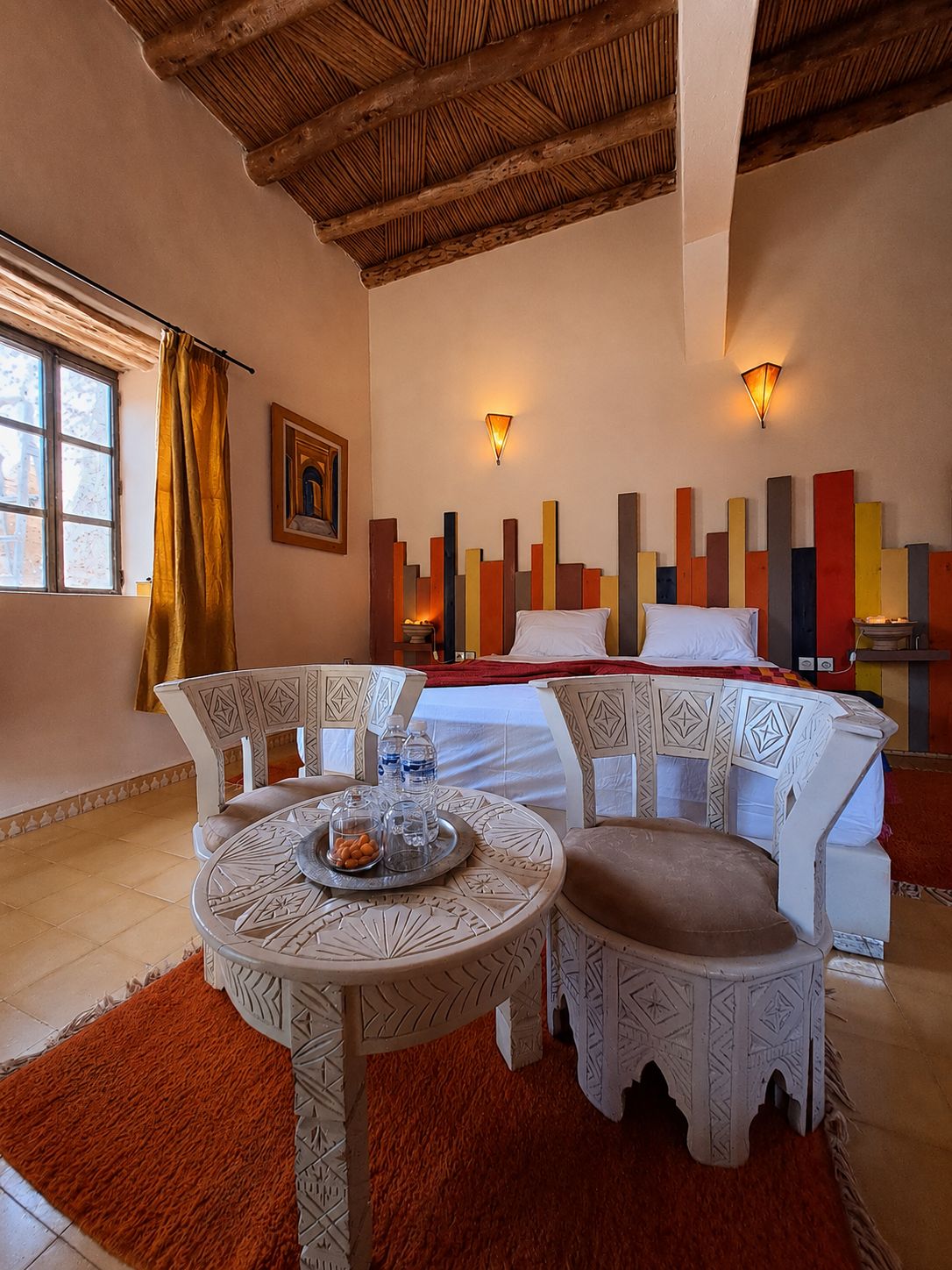 A hotel room with a bed, a dining table with chairs, a window with curtains, and a decorative wall panel.