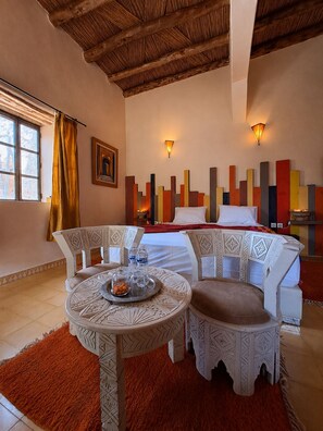 Una habitación de hotel con cama, una mesa de comedor con sillas, un ventanal con cortinas y un panel decorativo en la pared.