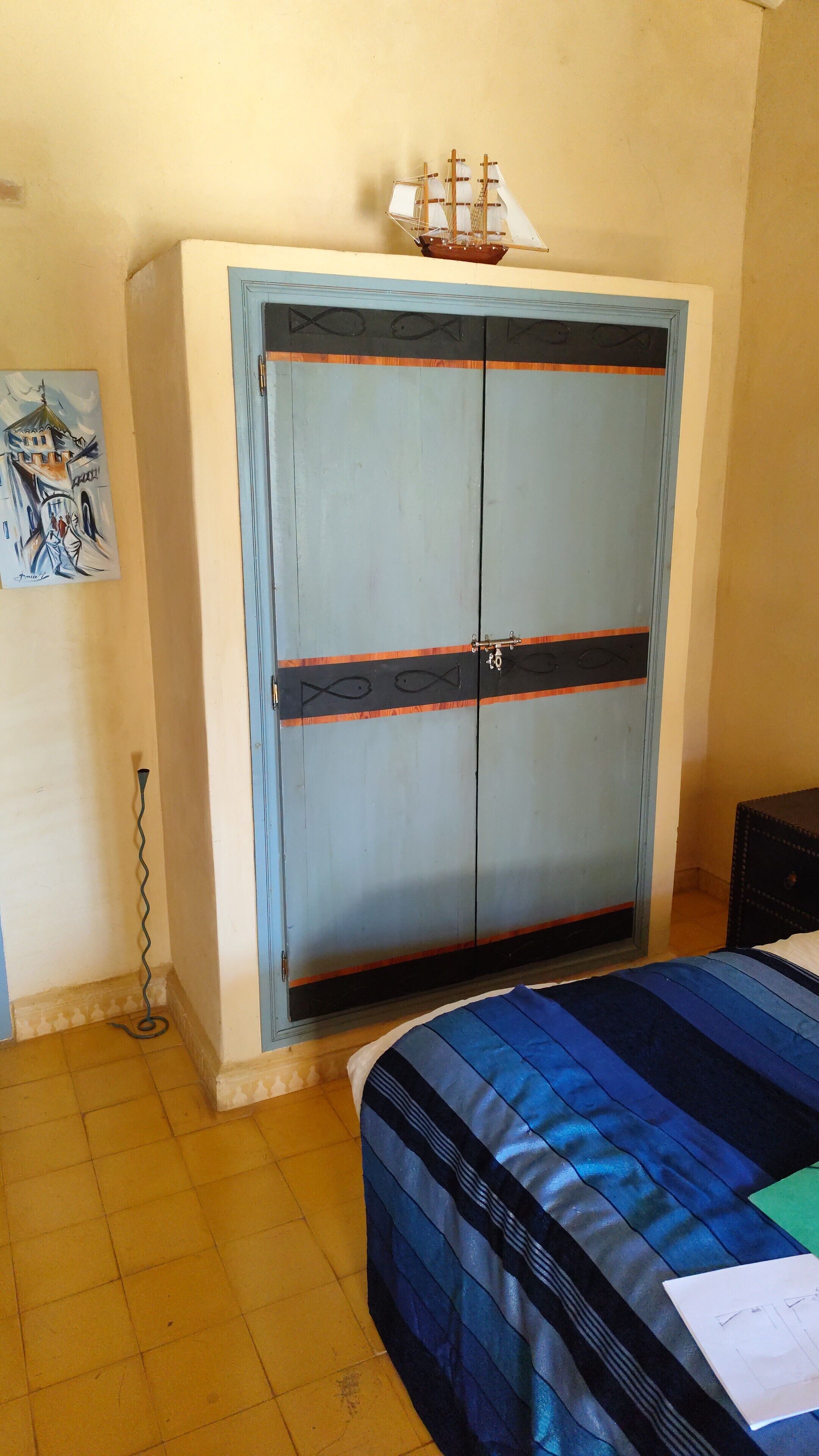 Una habitación con un armario grande, una cama con cubrecama azul y negro a rayas, y un pequeño modelo de barco encima del armario.