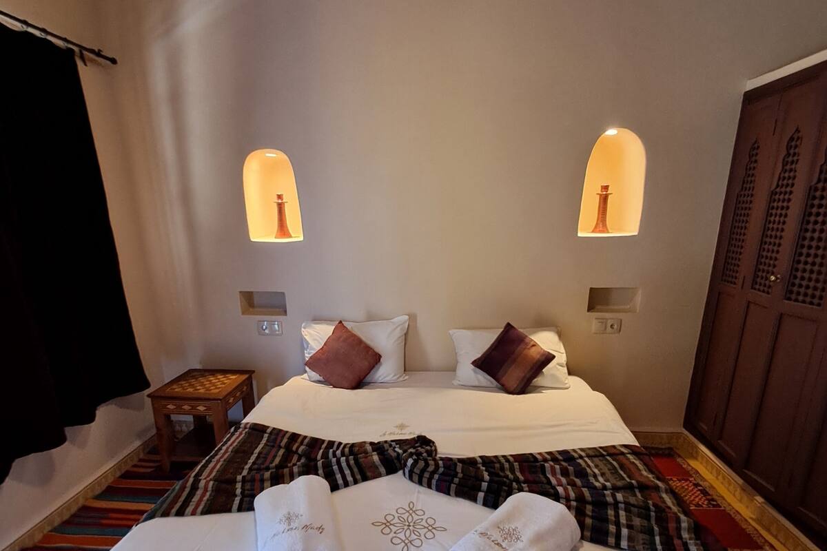 A hotel room with a large bed, two bedside lamps, a wooden wardrobe, and a small table with a chessboard.