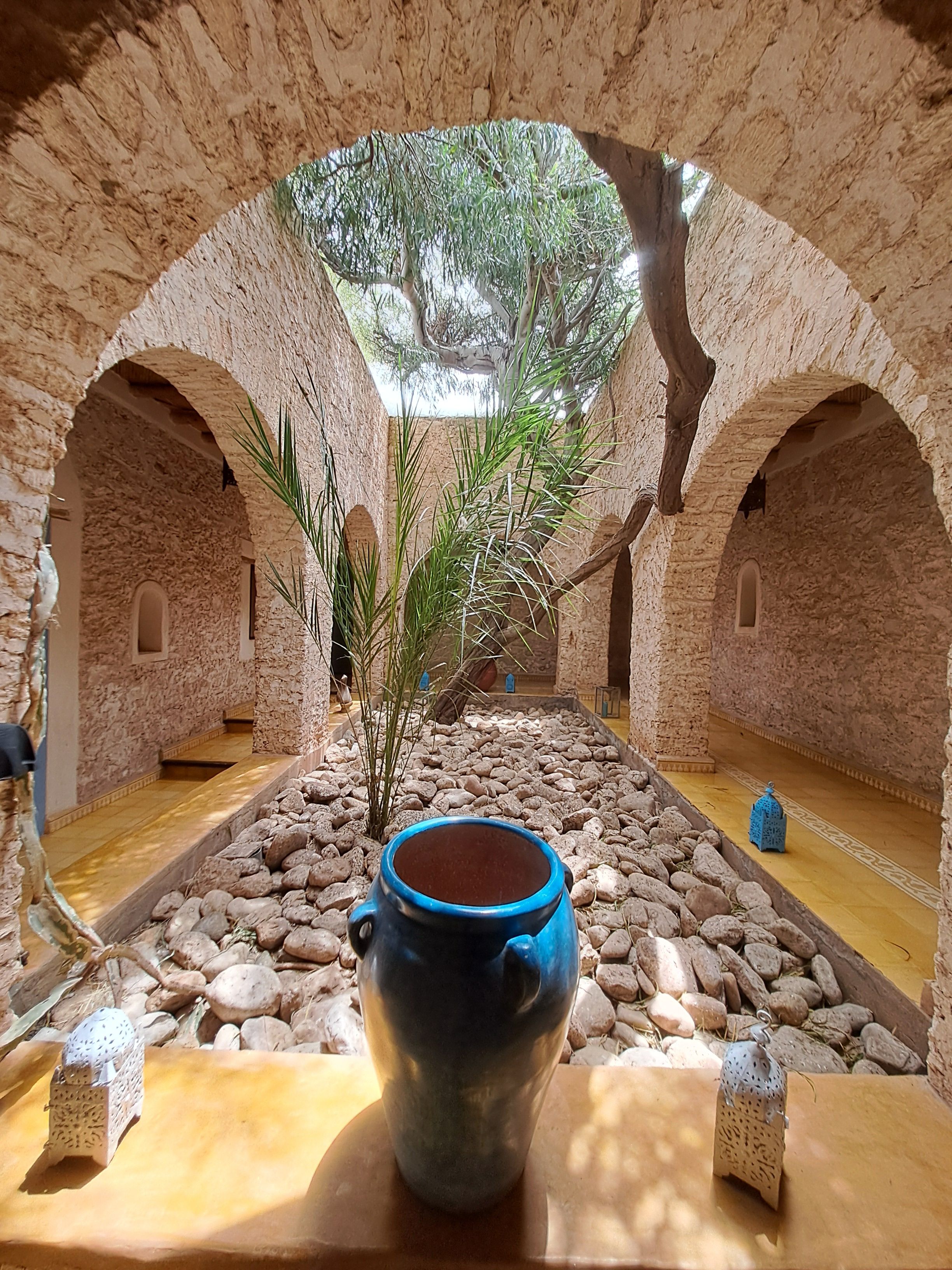 Un patio interior con arcos de piedra, una fuente central y una maceta azul sobre una mesa.