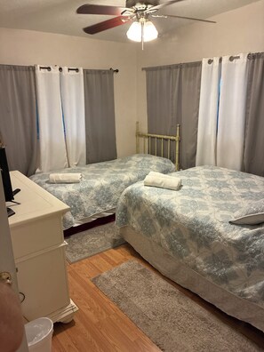 Ett sovrum med två sängar, ett skrivbord, en takfläkt och ett fönster med gardiner.