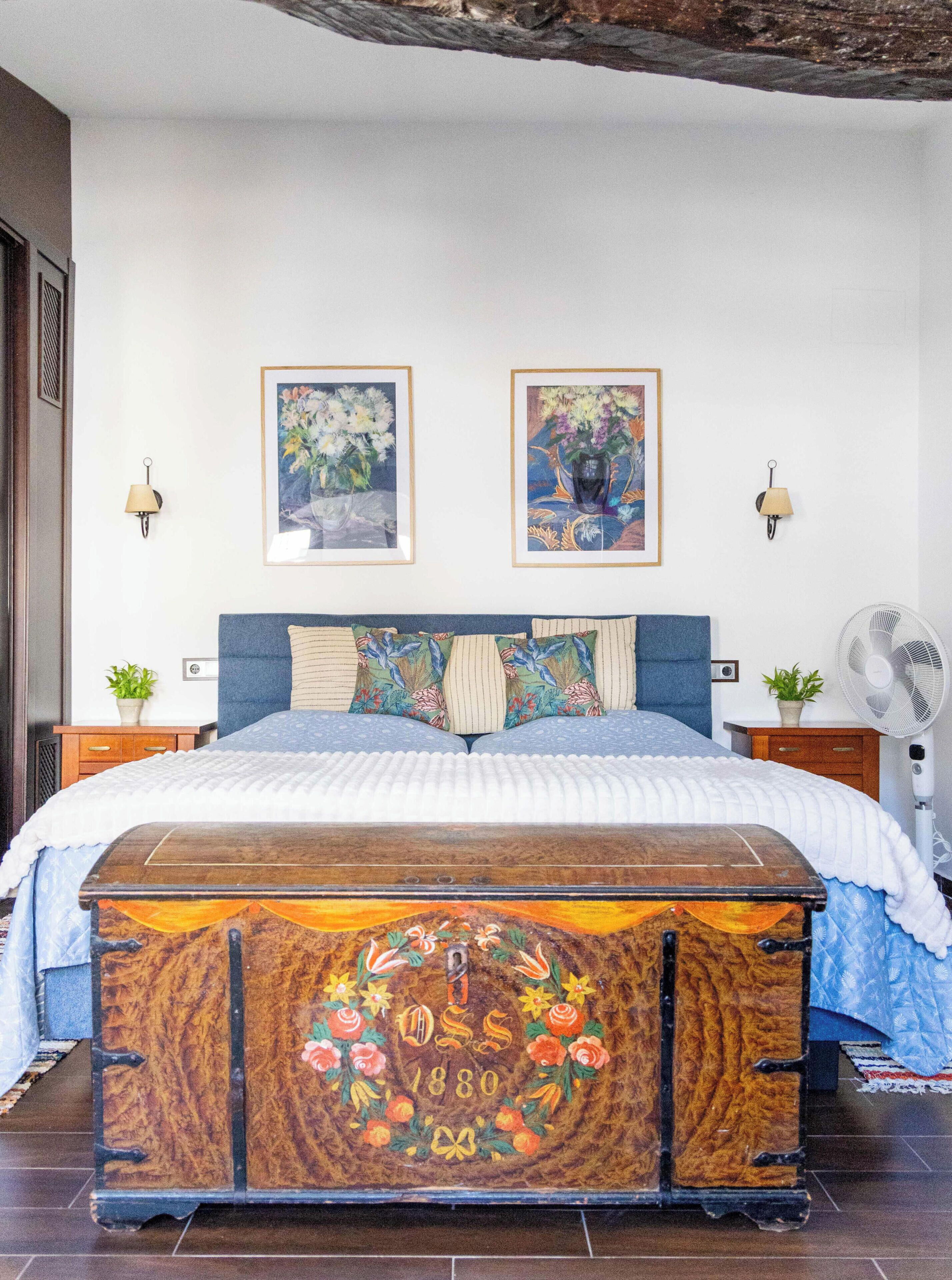 Een slaapkamer met een groot bed, twee wandlampen, een nachtkastje met een plant en een vintage koffer met bloemenmotieven.