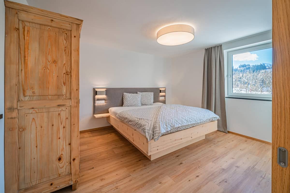 Ein Schlafzimmer mit Bett, Schrank, Nachttisch, Wandleuchte und Fenster mit Blick.