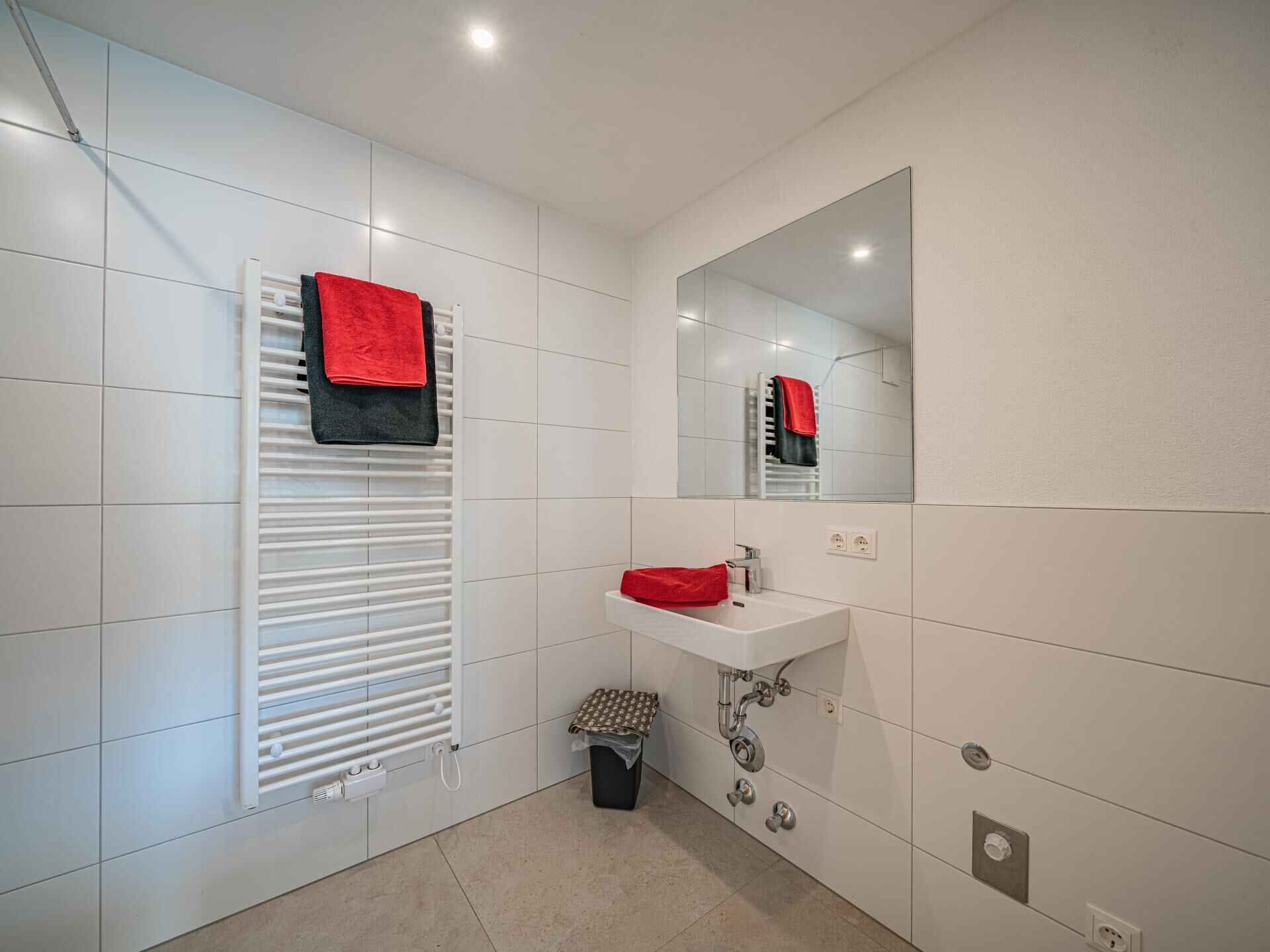 Een moderne badkamer met witte tegels, een witte radiator, een wastafel met een rode handdoek en een spiegel.