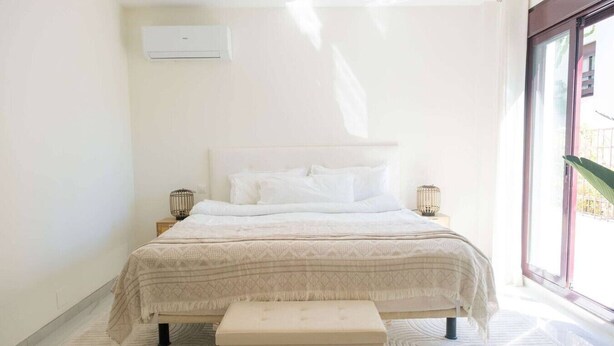 A bedroom with a bed, bedside tables, a bench, a window, and an air conditioning unit. - Modern 3 bed apt, walking distance to Puerto Banus (Marbella)