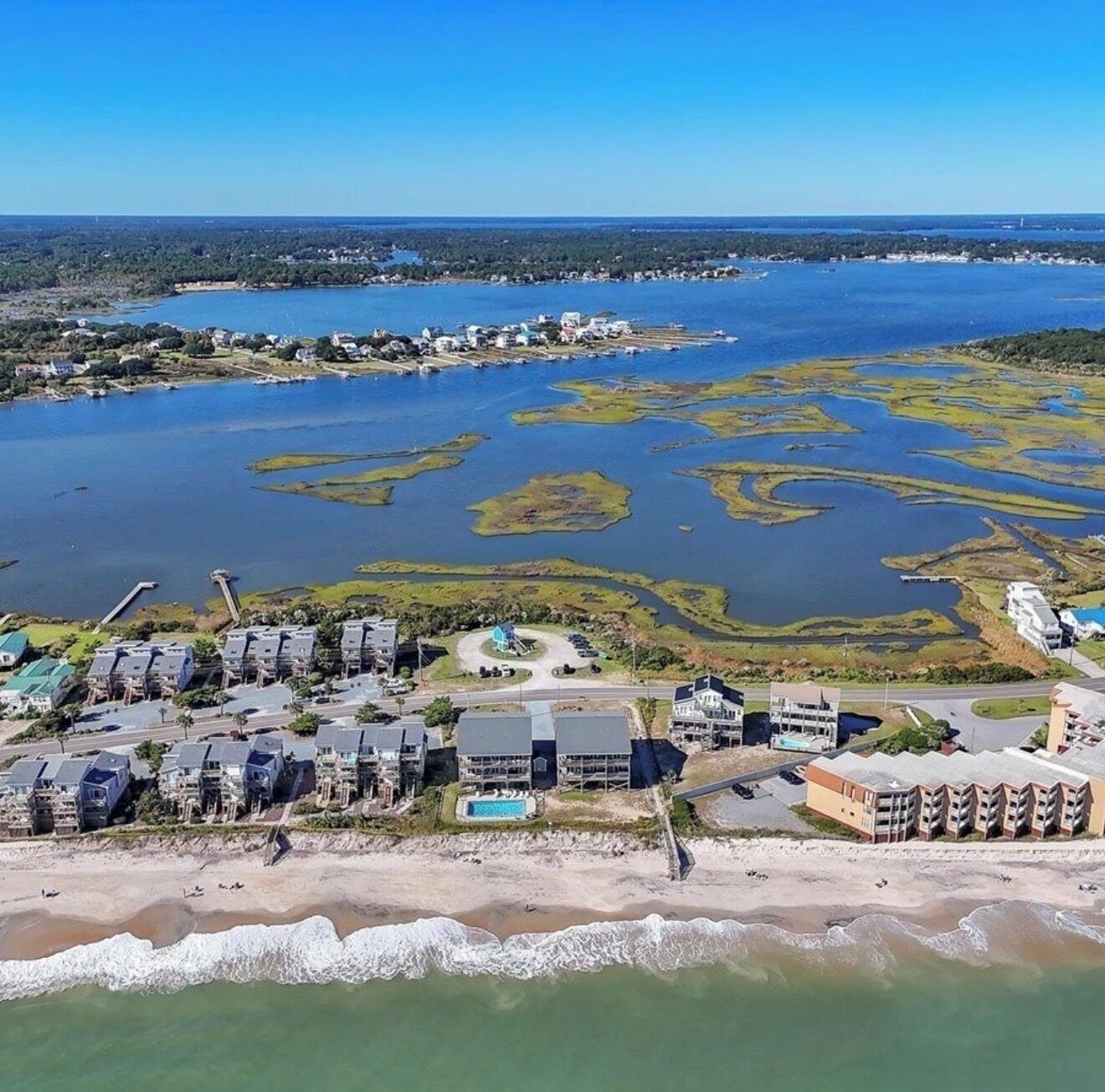 An aerial view of a coastal area with residential buildings, a beach, and a large body of water with marshy islands.