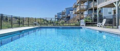 A swimming pool with a clear blue water surface, surrounded by a black fence, and adjacent to a multi-story building with balconies.