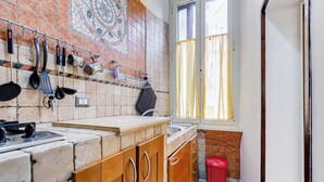 Eine Küche mit gefliestem Spritzschutz, hängendem Kochgeschirr, einem Fenster mit Vorhängen und einem roten Mülleimer.