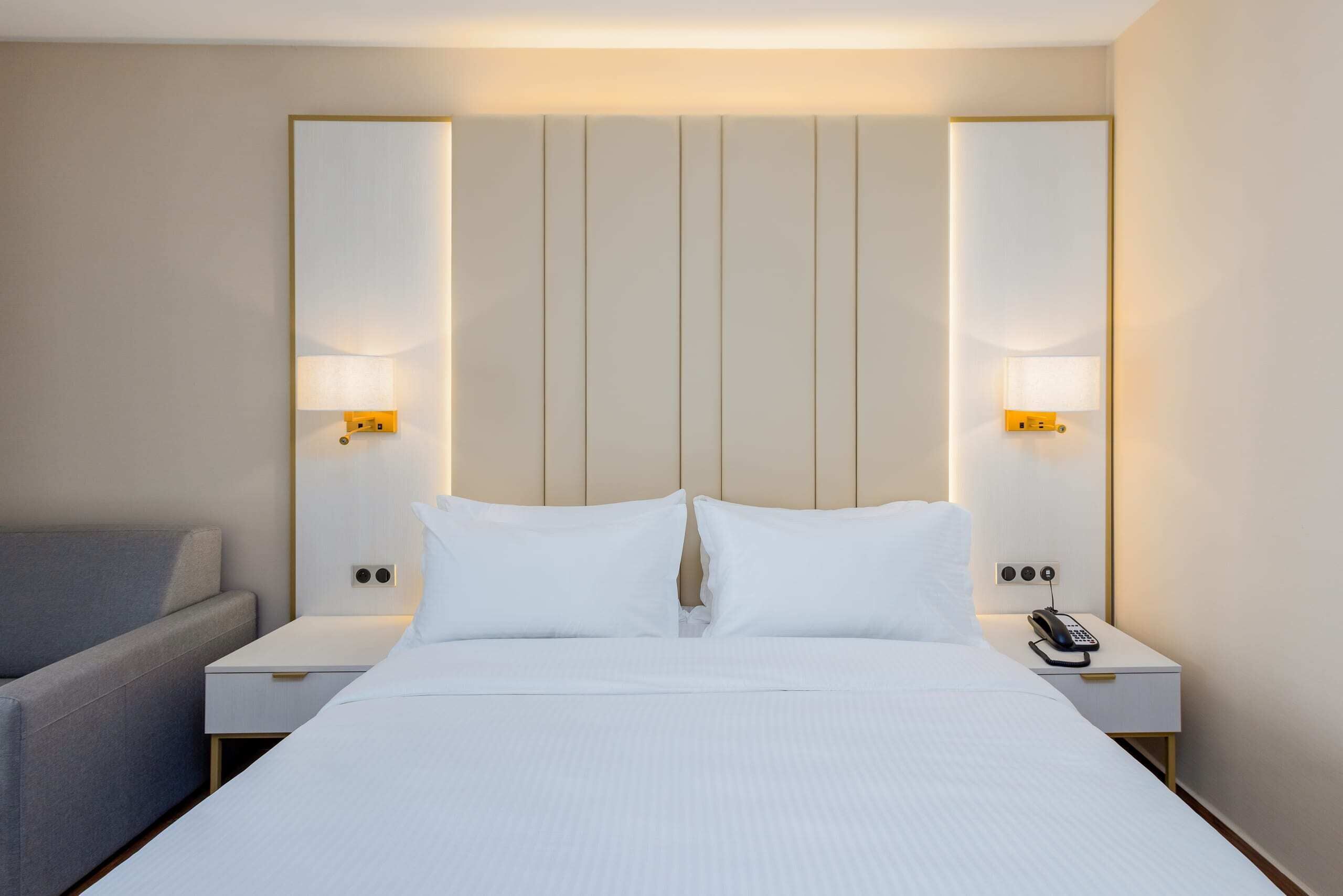 A hotel room with a large bed, two bedside tables, a bedside lamp, a phone, and a grey armchair.