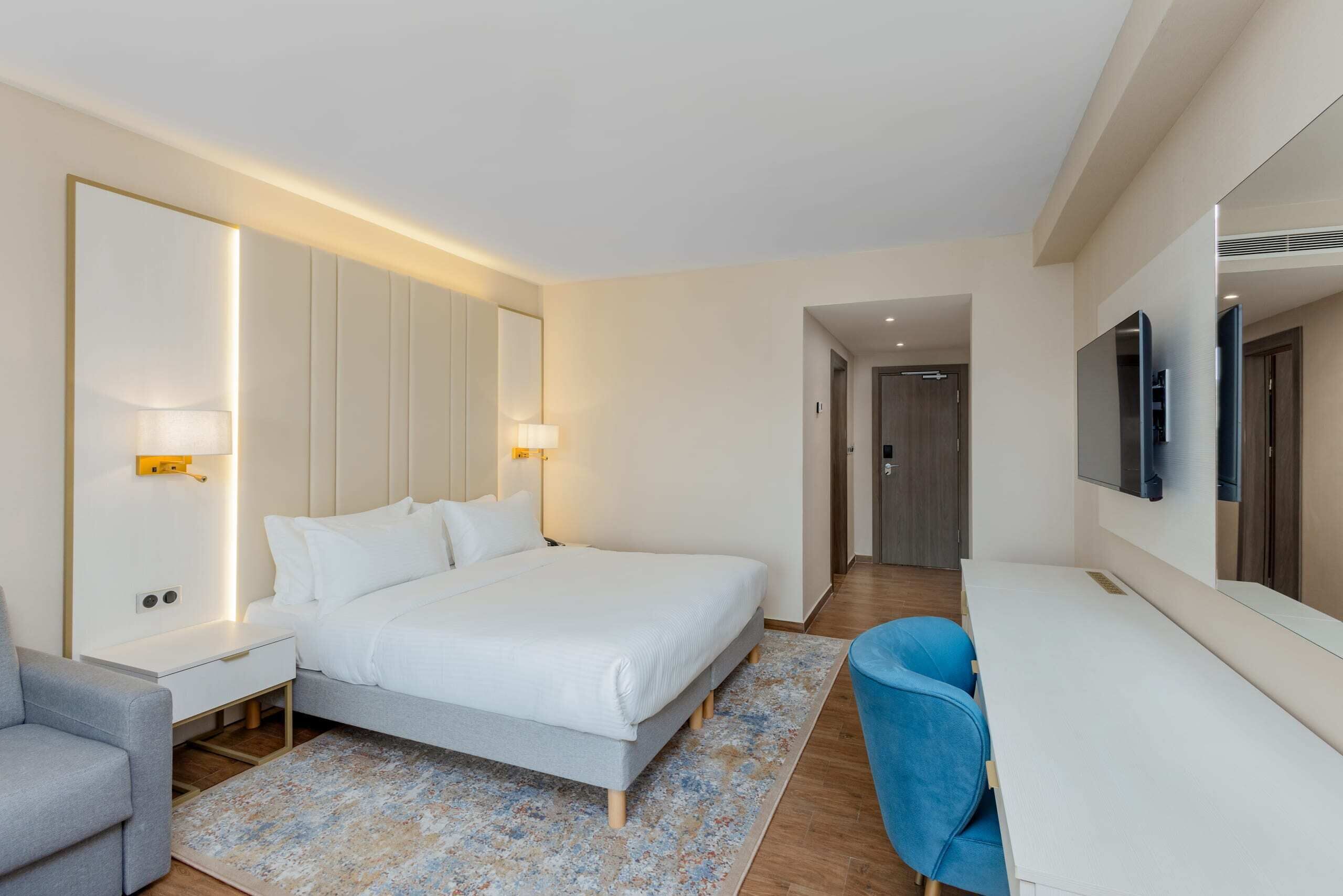 A modern hotel room with a large bed, bedside tables, a desk, a chair, and a TV.
