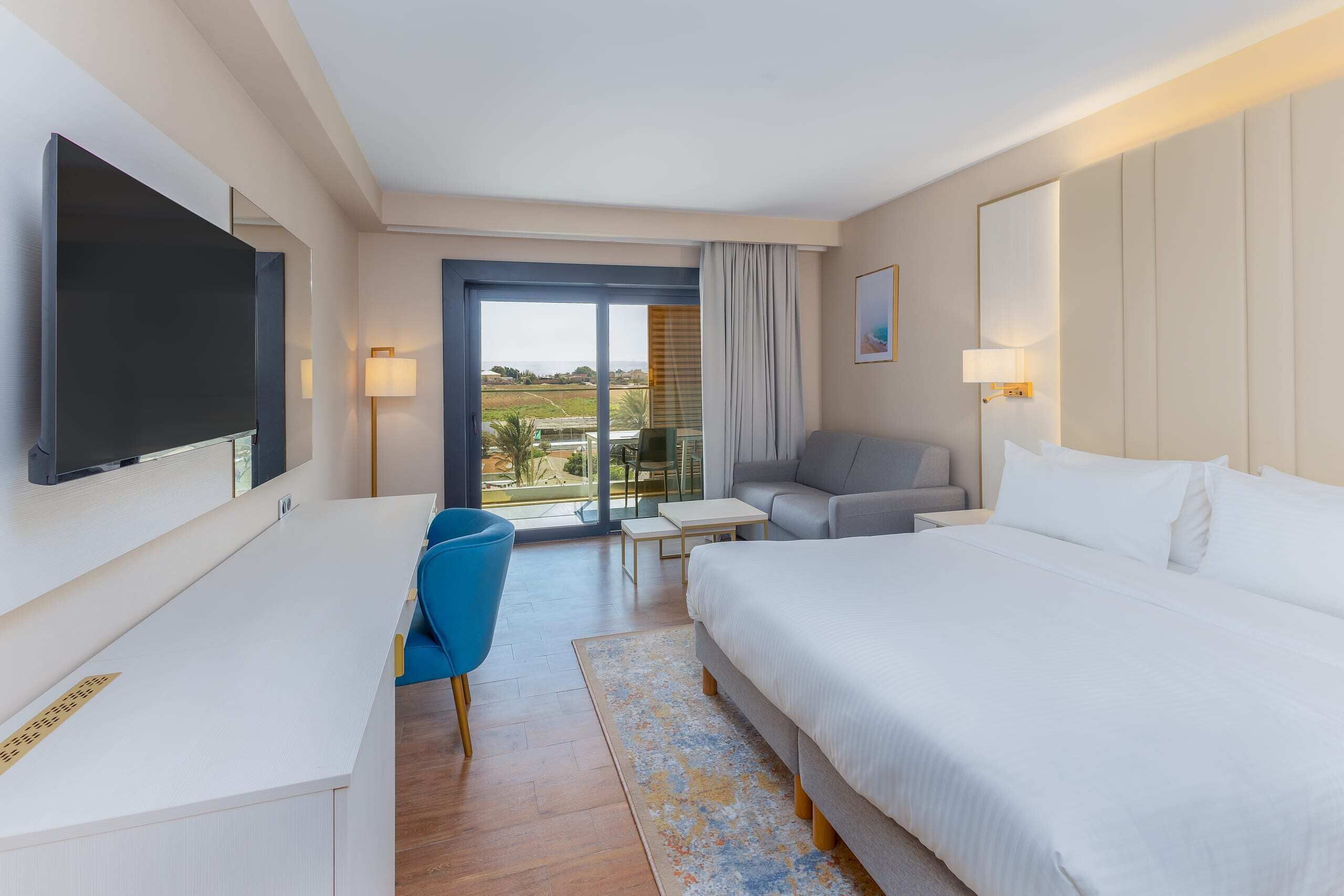 A hotel room with a bed, a desk, a chair, a TV, and a view of the outdoors.