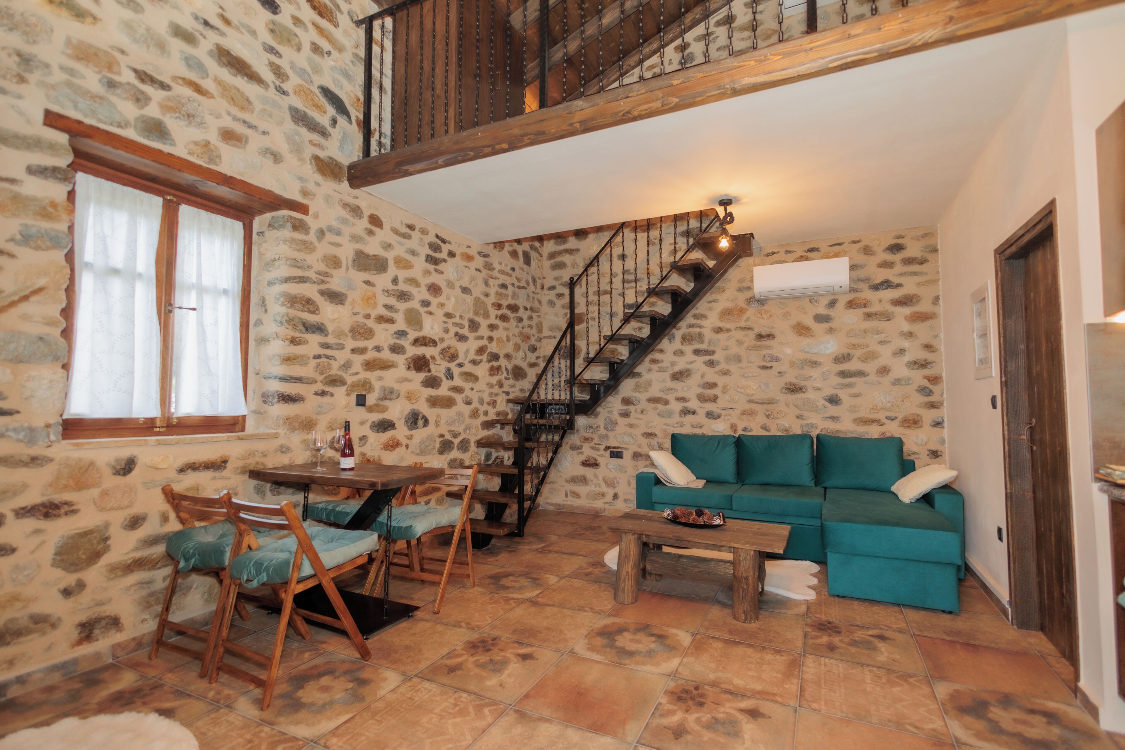 A stone-walled room with a staircase, a teal sofa, a wooden dining table, and chairs.