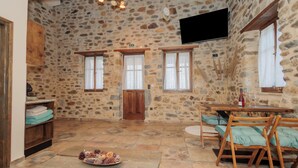 Una habitación con paredes de piedra, una puerta de madera, dos ventanas con cortinas, un televisor montado en la pared, una mesa redonda con sillas y una cesta de piñas en el suelo.