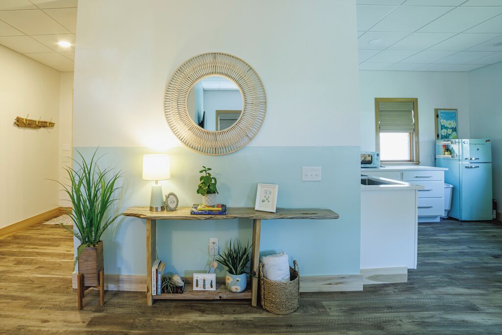 A hotel lobby with a reception desk, a round mirror, a lamp, potted plants, and a kitchenette in the background.