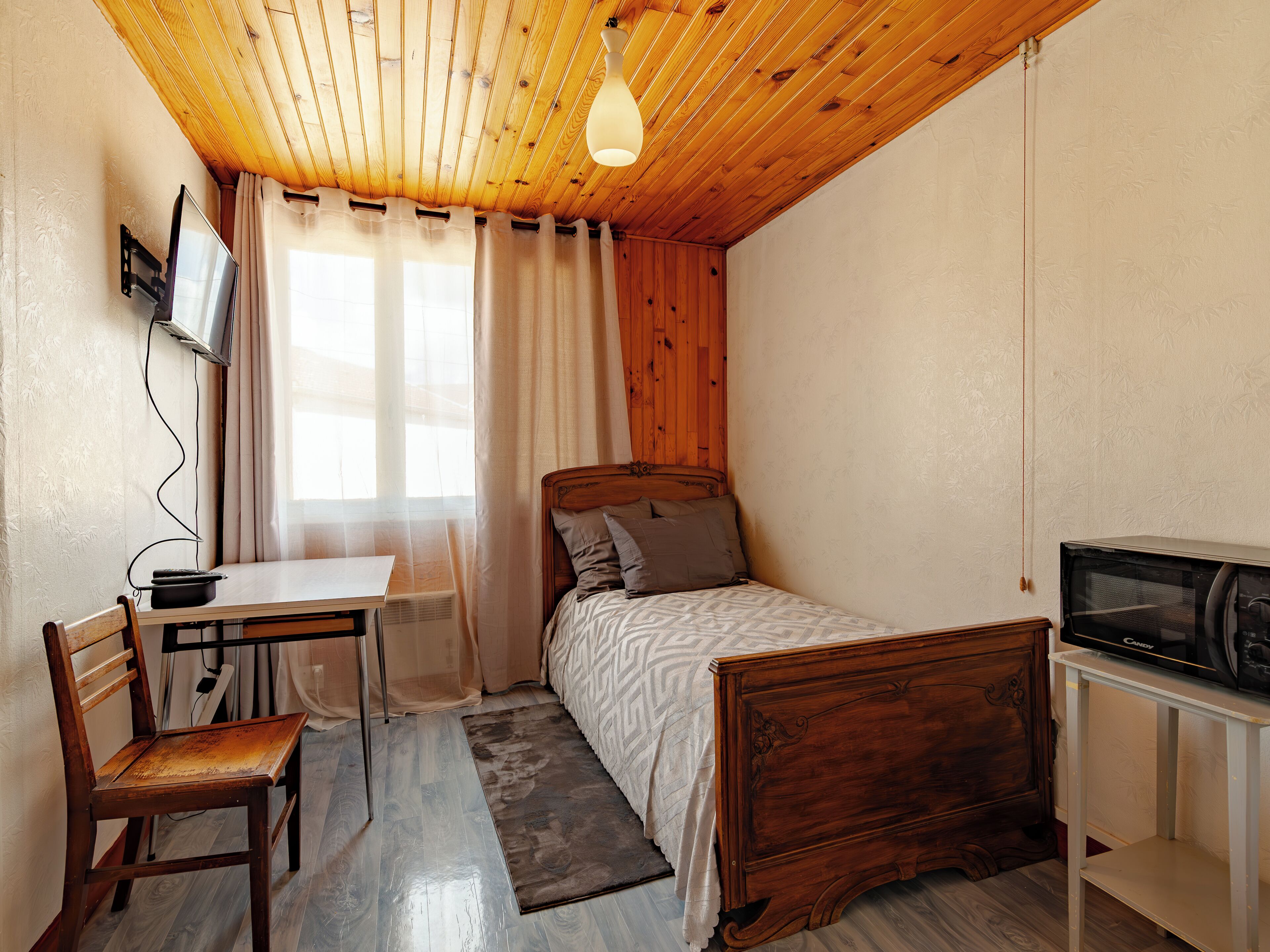 Ein kleines Schlafzimmer mit Holzboden, einem Bett, einem Schreibtisch mit Stuhl, einem Fernseher und einem Fenster mit Vorhängen.