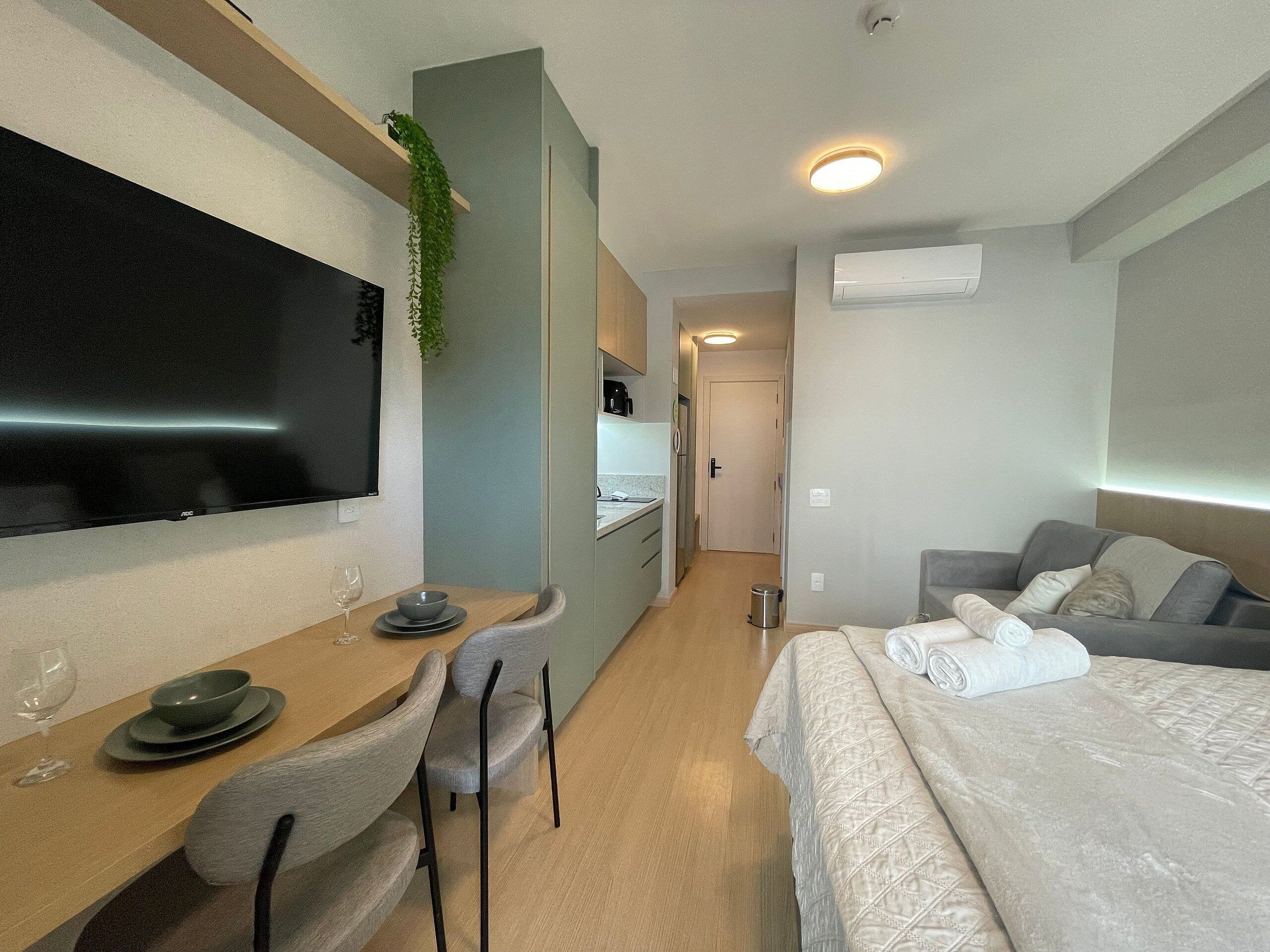 A hotel room with a bed, a desk with a chair, a TV, a plant, and a kitchenette.