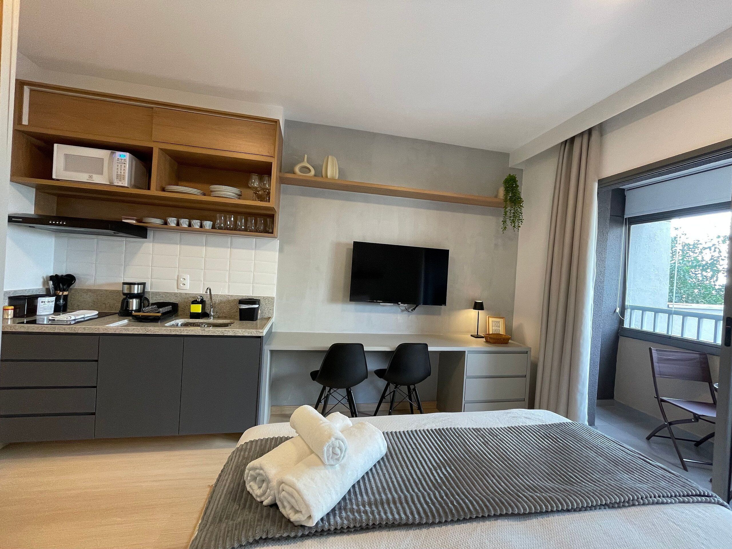 A modern hotel room with a kitchenette, a bed with a grey textured bedspread, a flat-screen TV, and a balcony with a view.