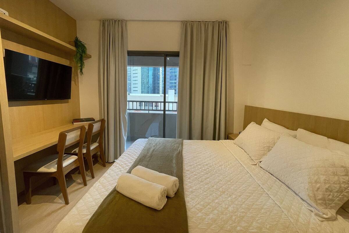 Quarto de hotel com cama, escrivaninha, cadeira, televisão e varanda com cortinas.